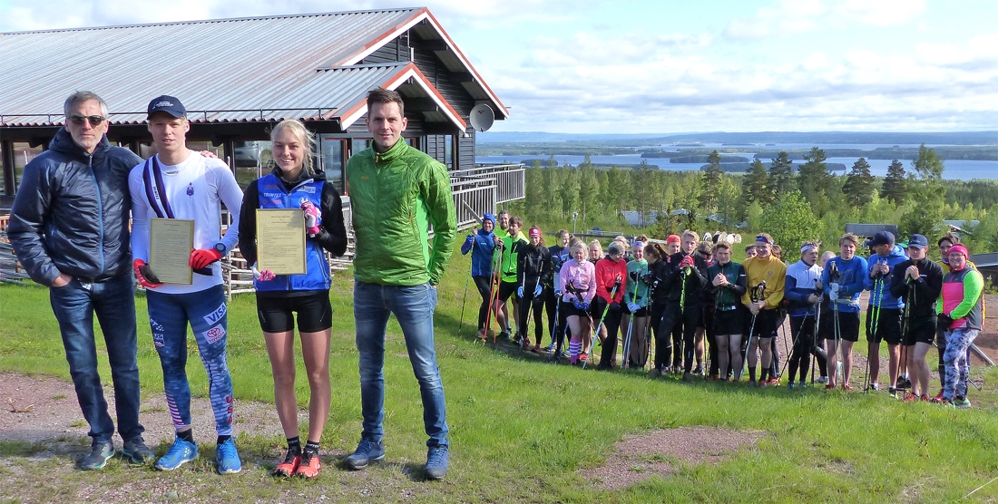 Erik Svensson fr&aring;n styrelsen f&ouml;r Mora Nisses Jubileumsfond och Mora Nisses barnbarn Erik Eriksson delade ut stipendierna till Axel Aflodal och Louise Lindstr&ouml;m. I bakgrunden elever och l&auml;rare vid Mora skidgymnasium. FOTO: Mora Nisses Jubileumsfond.